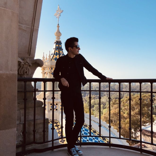 Peter, a dark-haired man dressed in dark clothing, leans against a metal railing overlooking a religious building with a mosaic roof and trees
